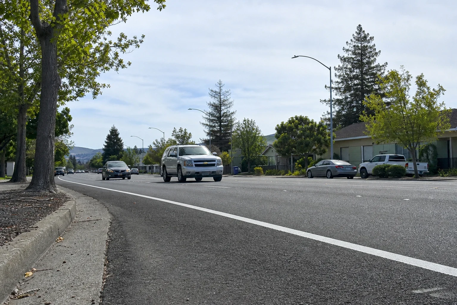 Low view of a wide suburban street with a silver SUV and cars in light traffic, tall pines, street lamps, single-story homes and hills under a high-cloud sky