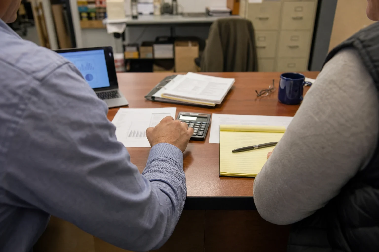 Two people at an office desk reviewing printed statements, one using a calculator beside a yellow notepad, laptop and coffee mug, with filing cabinets behind