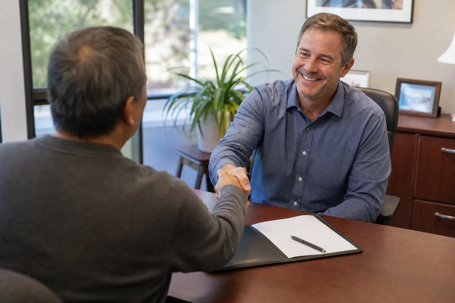 Smiling agent in a blue shirt shakes hands with a client across a wooden office desk with an open folder and pen, a plant and window behind