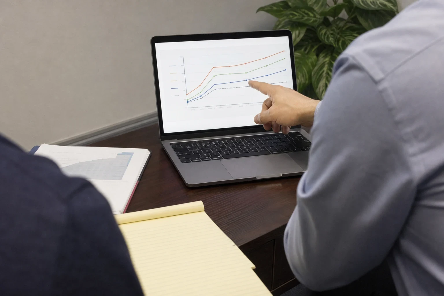 Over-the-shoulder view of an agent pointing at a line chart on a laptop for a client, with a printed document and yellow legal pad on a dark wood desk beside a plant
